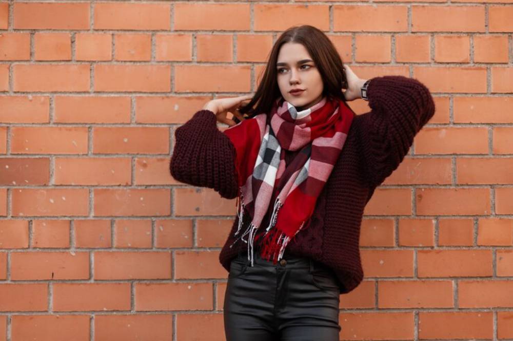 A person poses against a brick wall, wearing a cozy maroon sweater and a red plaid scarf, with hands in hair and a confident stance.