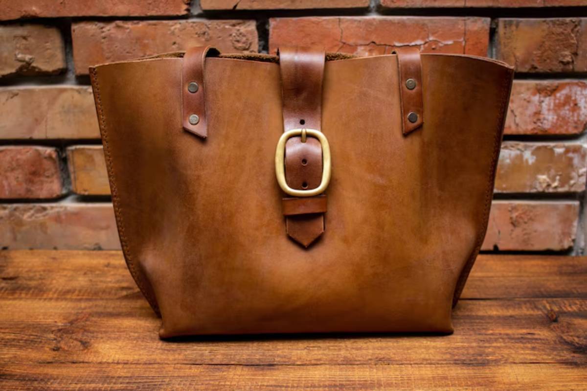 A stylish brown leather tote bag with a large belt buckle and straps, set against a rustic brick background.