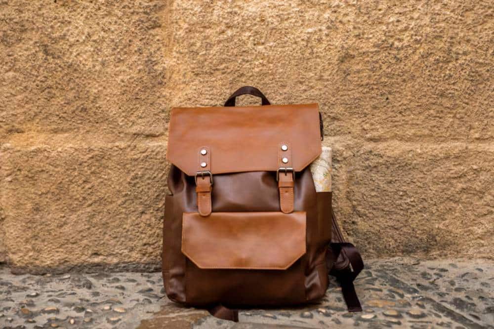 Stylish brown cork leather backpack with a front pocket, set against a textured stone wall. A map peeks out from the side.