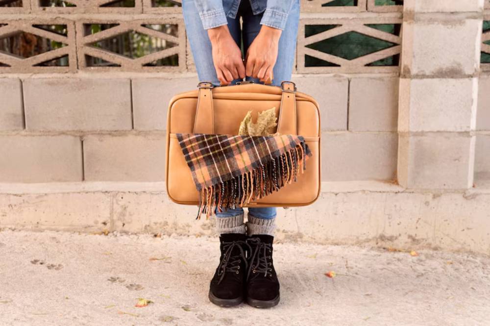 A person holds a tan colored bag with a plaid scarf draped over it, wearing denim and black boots, standing against a textured background.