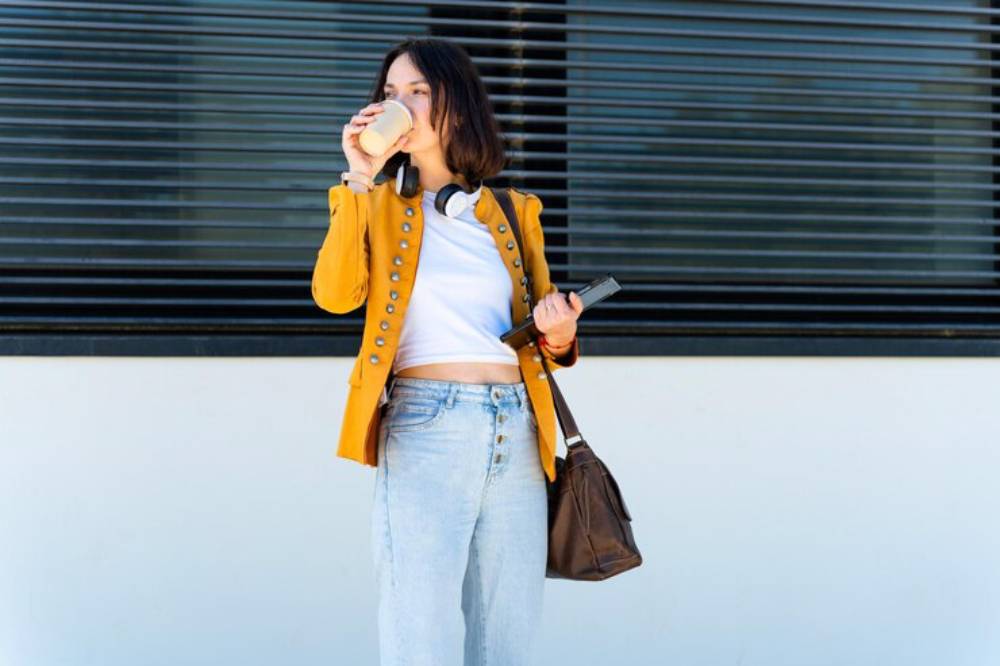 A person stands against a building, wearing a yellow jacket and light jeans, holding a phone and bag, with headphones around their neck.