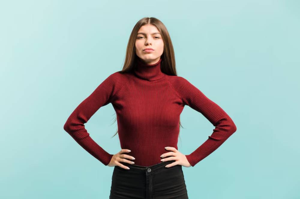 A person in a fitted burgundy turtleneck and black pants stands confidently, hands on hips, against a light blue background.