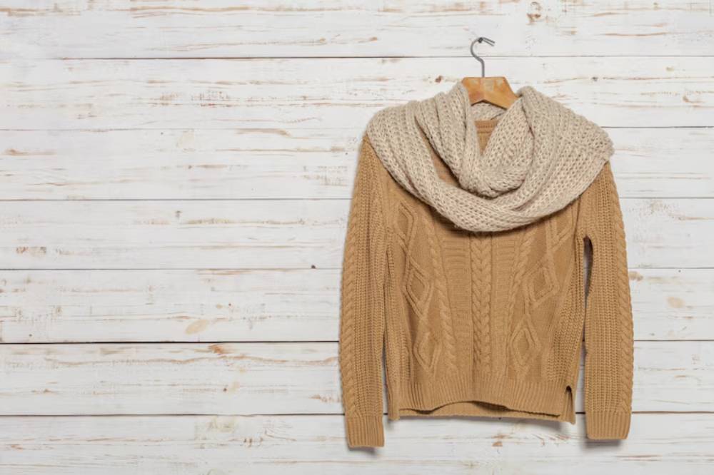 A cozy tan cable-knit sweater draped with a light beige scarf, hanging on a wooden wall.