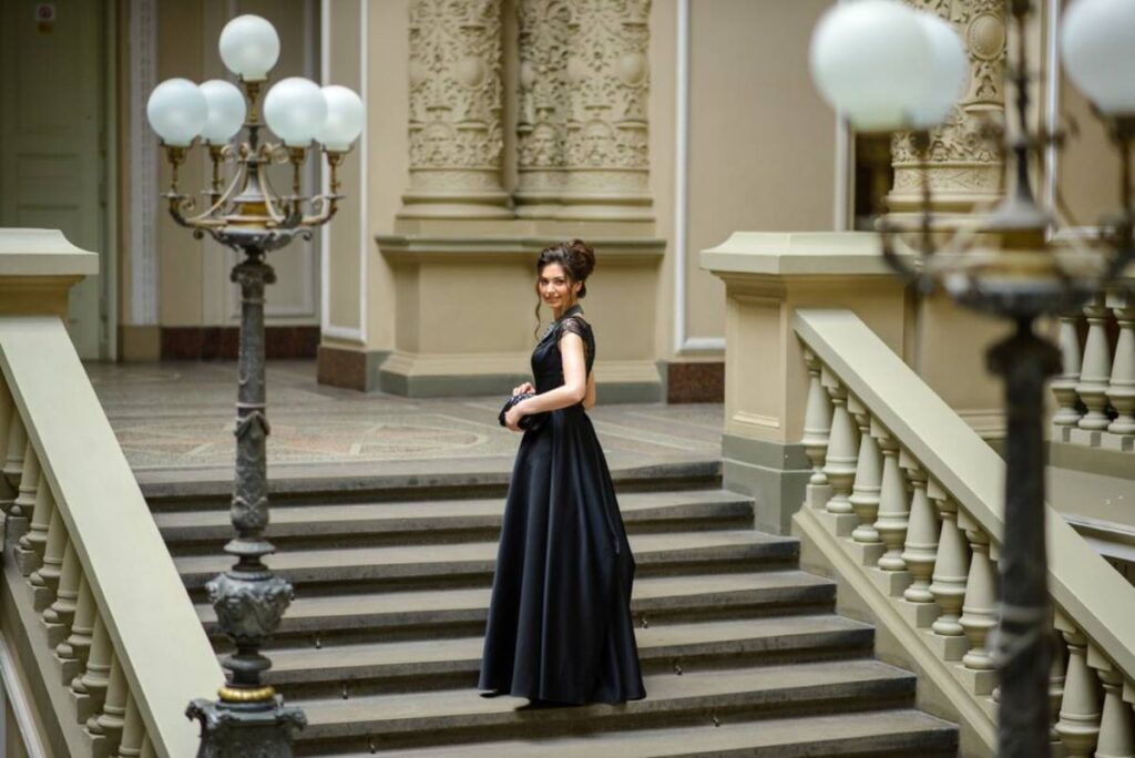 A woman stands gracefully on ornate stairs in a long black gown, surrounded by elegant architectural details and vintage lamps.