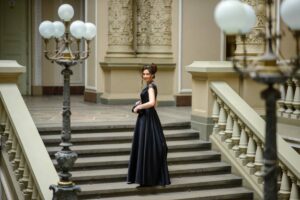 A woman stands gracefully on ornate stairs in a long black gown, surrounded by elegant architectural details and vintage lamps.