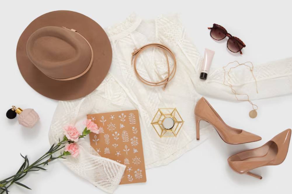 A flat lay of fashion accessories including a brown hat, nude heels, a white blouse, sunglasses, and a floral notebook, accented with flowers.