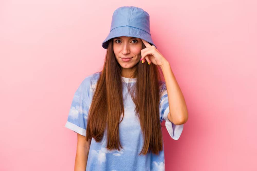 A young woman poses in front of a pink background, wearing a sky-blue bucket hat and a cloud-patterned t-shirt, with long, straight hair.