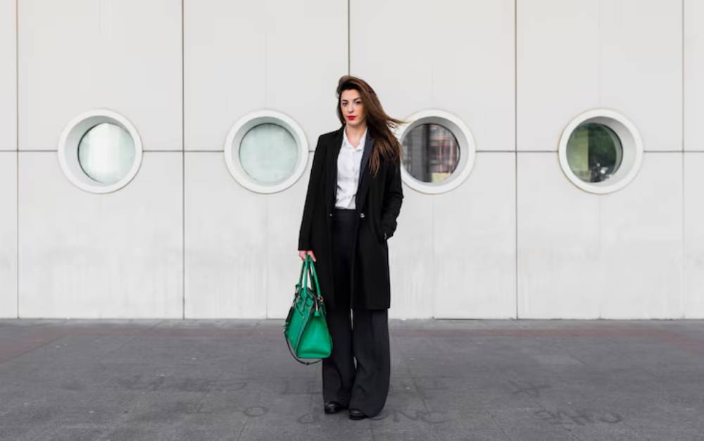 A woman in stylish attire stands confidently in front of a modern, white wall with circular windows, holding a green handbag.
