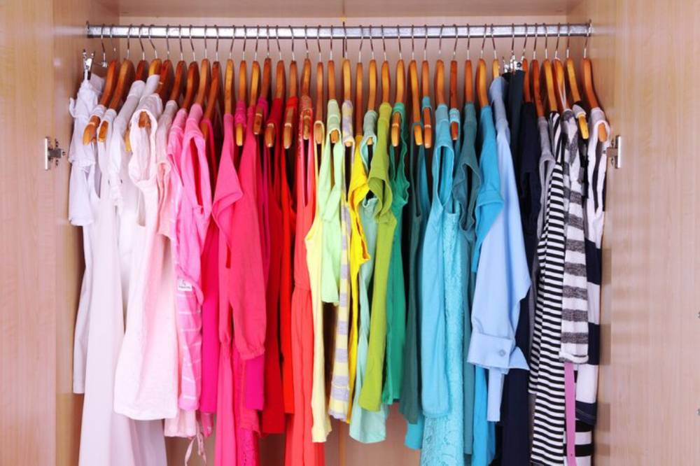 A colorful array of clothing hanging neatly on wooden hangers inside a light wood closet. The garments include various styles and colors.
