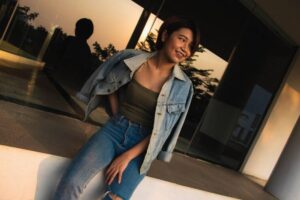 A person sits casually on a ledge, wearing a denim jacket and high-waisted jeans, as the sunset casts a warm glow.