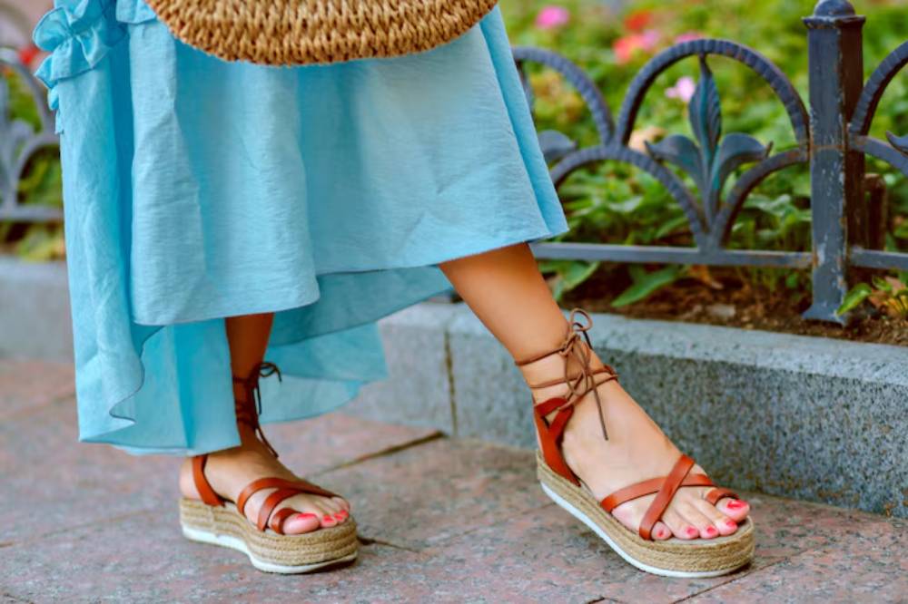 A close-up of a woman’s feet in brown strappy espadrilles, paired with a flowing blue dress and a woven bag, set against colorful flowers.