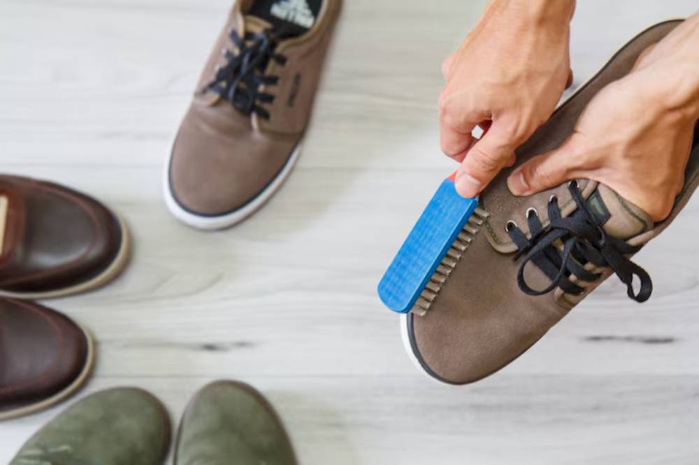 A person cleans a brown shoe using a blue shoe brush, with several other shoes visible on a wooden floor.
