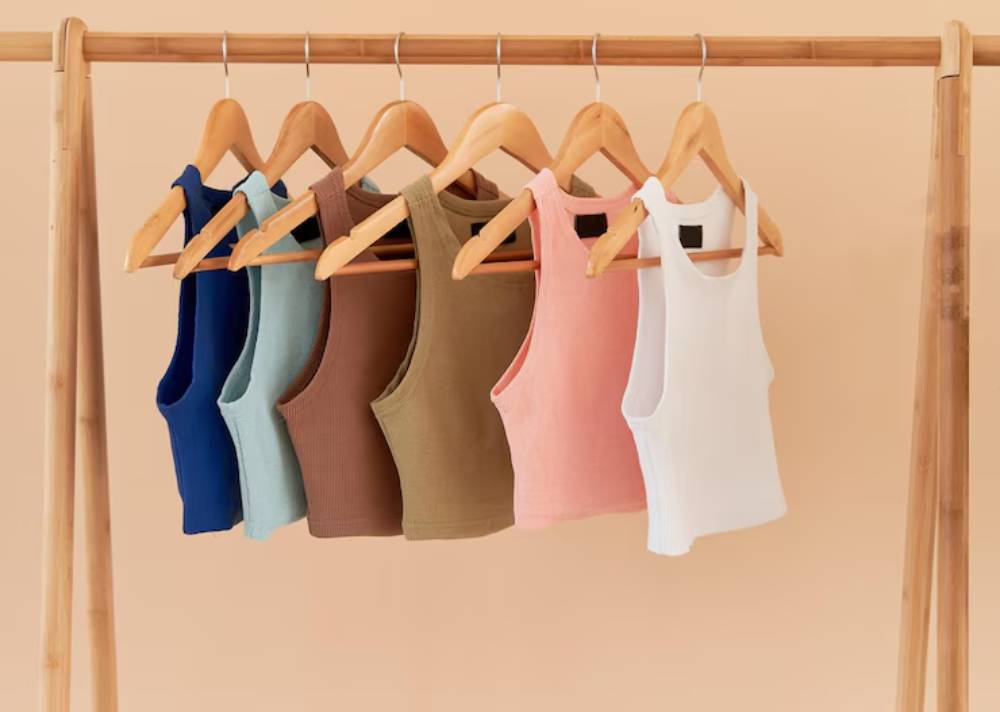 A row of six colorful tank tops hanging on wooden hangers against a soft peach background.