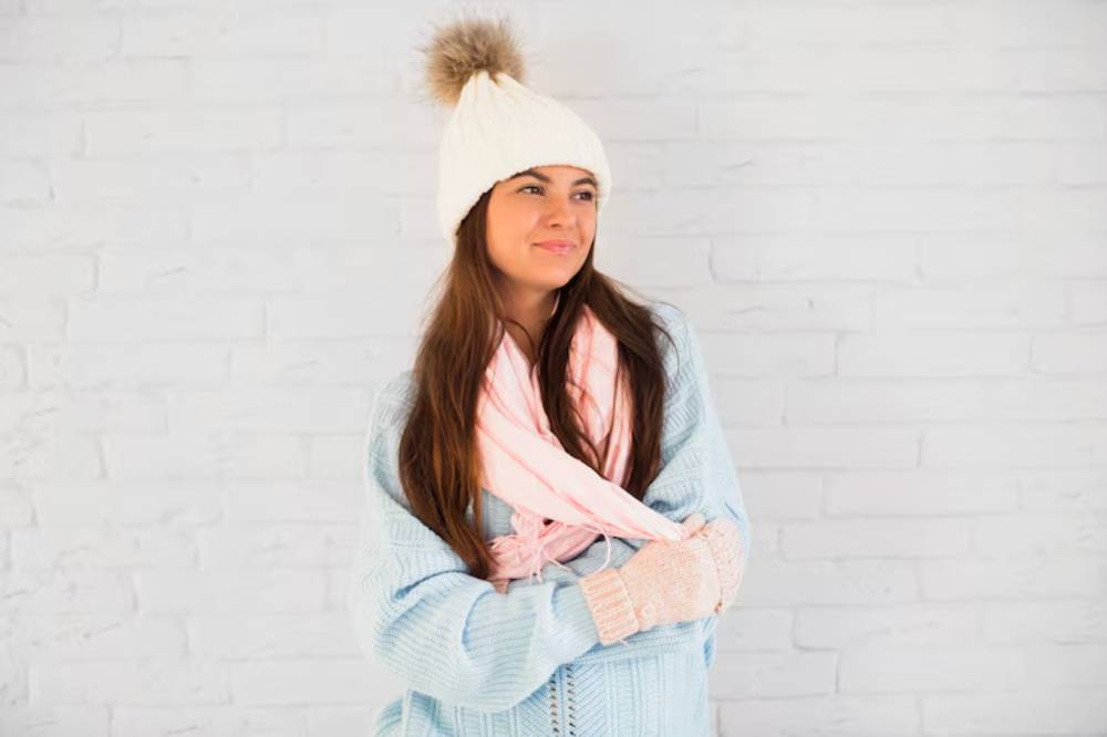 A woman in a cozy light blue sweater, cream beanie, and pink scarf stands against a white brick wall, crossing her arms.