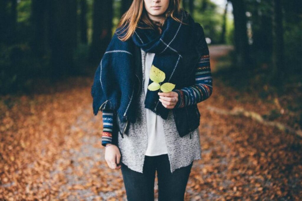 A woman stands on a leaf-strewn path, holding green leaves, dressed in a cozy navy scarf and a multicolored sweater.
