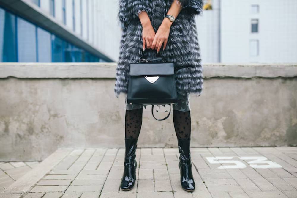A woman in a furry coat, black bag, and glossy boots stands confidently, showcasing a stylish urban look against a modern backdrop.