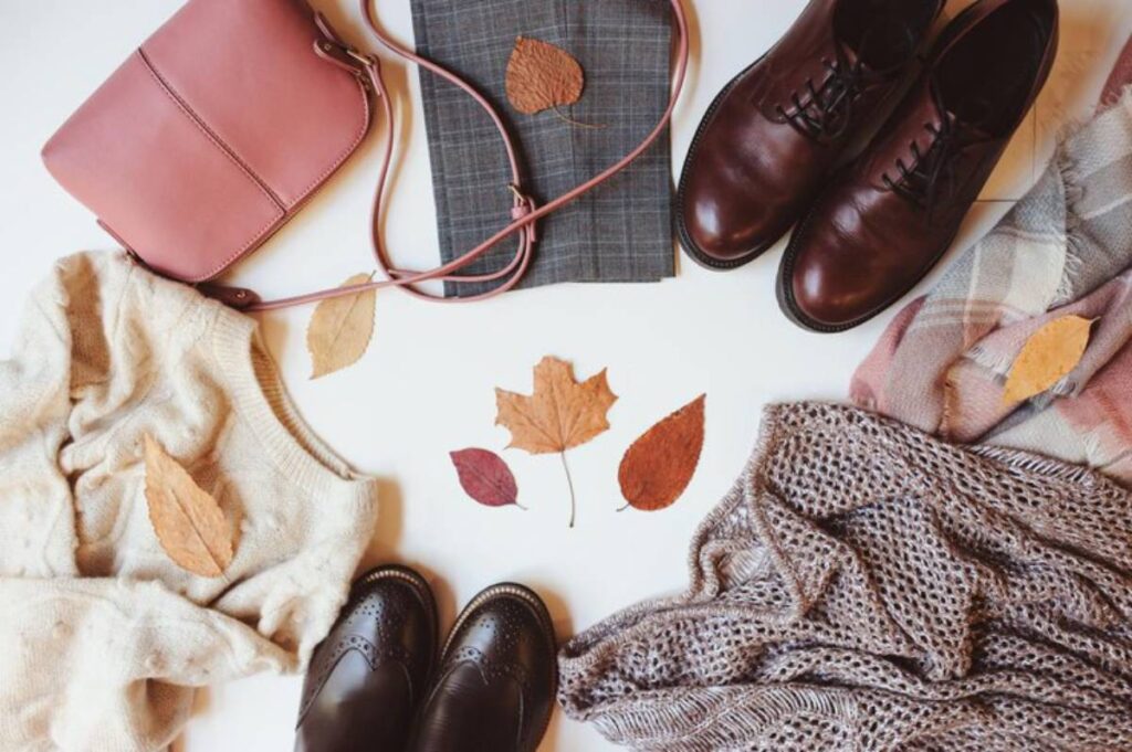 Flat lay of cozy fall outfits featuring a beige sweater, brown shoes, a pink handbag, plaid fabric, and autumn leaves scattered around.