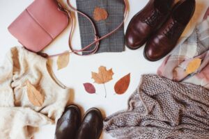 Flat lay of cozy fall outfits featuring a beige sweater, brown shoes, a pink handbag, plaid fabric, and autumn leaves scattered around.