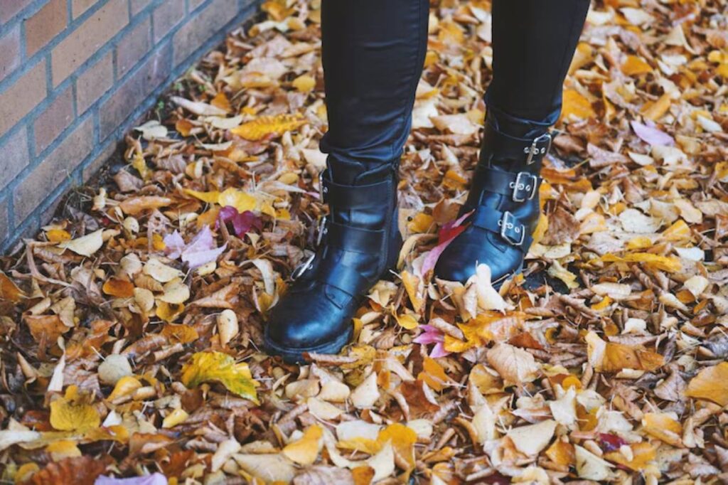 A close-up of black buckle boots standing on a colorful carpet of autumn leaves, with a brick wall in the background.