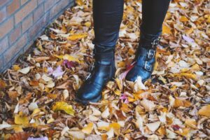 A close-up of black buckle boots standing on a colorful carpet of autumn leaves, with a brick wall in the background.