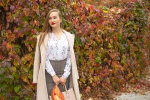 A person in a floral blouse and plaid skirt stands against a backdrop of vibrant fall foliage, holding a beige handbag and an orange umbrella.