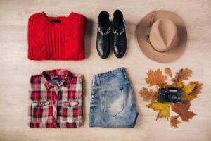 A flat lay of autumn apparel: a red sweater, plaid shirt, jeans, black boots, a hat, fall leaves, and a vintage camera.