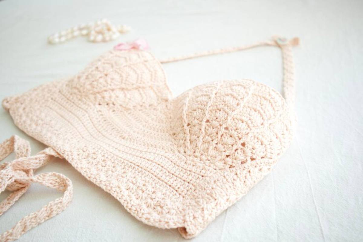 A handmade crocheted bikini top in soft pink, featuring textured patterns and ties, laid flat on a light surface with a pearl necklace nearby.