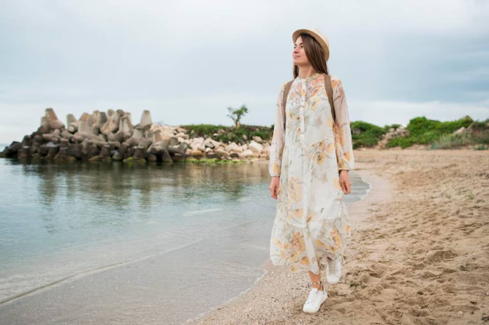 A woman in a floral dress and white sneakers walks along a sandy beach, with rocky formations and greenery in the background.