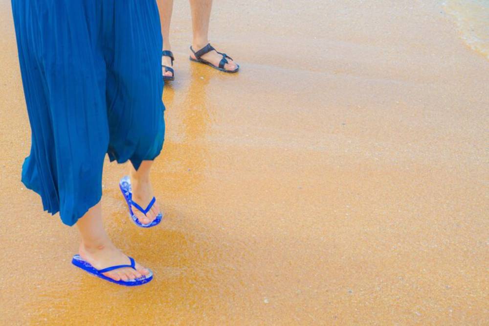 A person in a flowing blue skirt walks barefoot on a sandy beach, while a companion in sandals stands nearby.