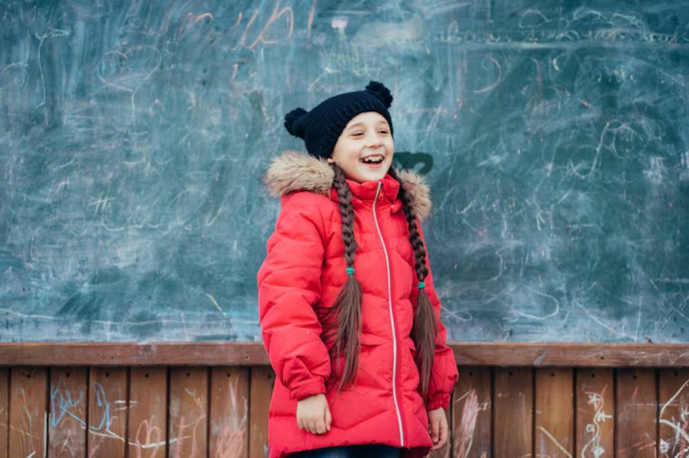 A child stands before a chalkboard, wearing a red winter coat with a fur-lined hood and a black beanie, showcasing long braided hair.