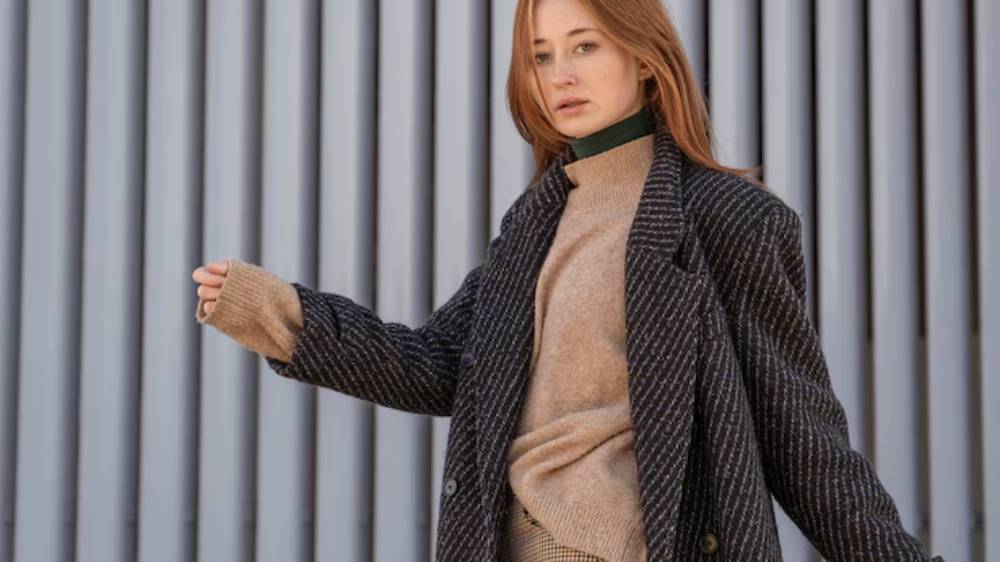 A person in a stylish layered outfit, featuring a dark, striped coat and a beige sweater, poses against a striped wall background.
