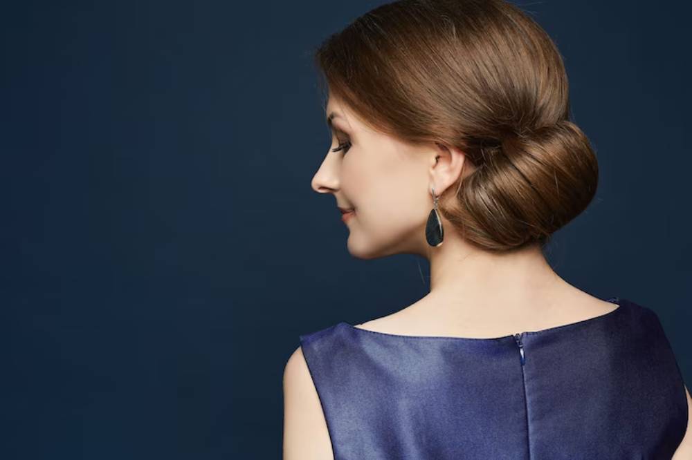 A woman with elegantly styled hair in a smooth bun, wearing a dark blue dress and statement earrings, poses against a dark background.