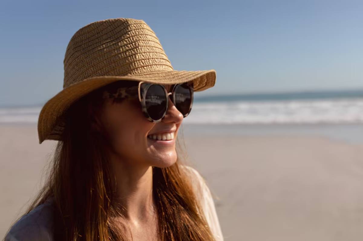 A woman with long hair wears a straw hat and sunglasses, standing on a sunny beach with waves in the background.