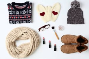 A cozy winter flat lay featuring a patterned sweater, scarf, mittens, hat, glasses, makeup brushes, lipsticks, and warm boots.