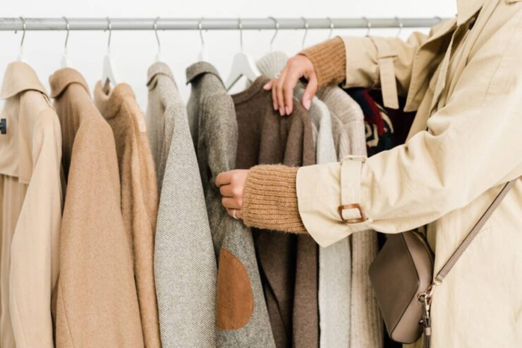 A person in a beige coat examines a rack of various stylish sweaters and coats in neutral tones, showcasing a cozy fashion selection.