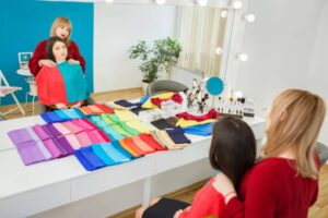 A brightly lit room featuring a makeup station with colorful fabric swatches neatly arranged on a table, and two women engaged in a discussion.