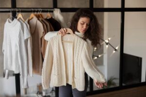 A person holds a cream-colored knitted cardigan, while several other sweaters hang on a rack in a cozy, modern setting.