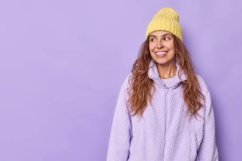 A person wearing a cozy lavender fleece jacket and a bright yellow beanie stands against a soft purple background.
