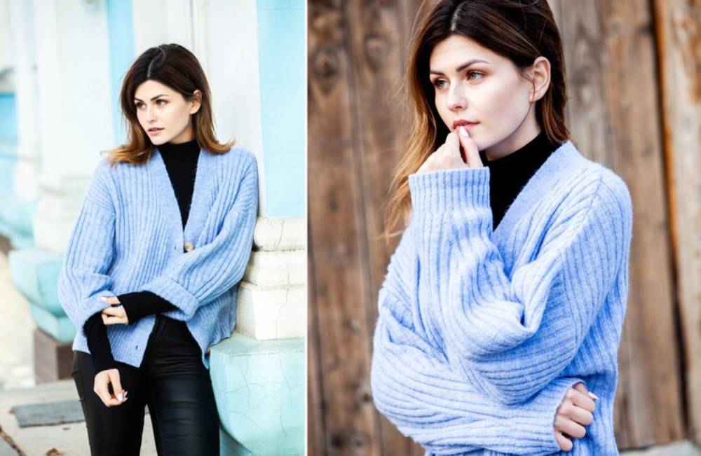 A person wearing a light blue oversized cardigan over a black turtleneck, standing against a rustic wooden backdrop.