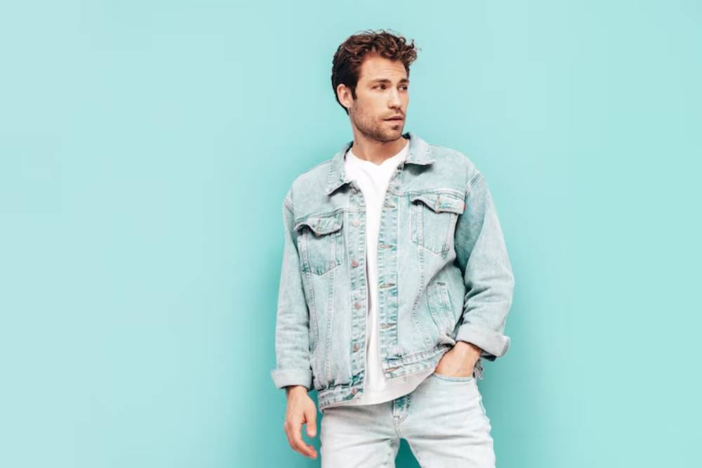 A person stands casually against a light blue background, wearing a distressed denim jacket, a white t-shirt, and light jeans.