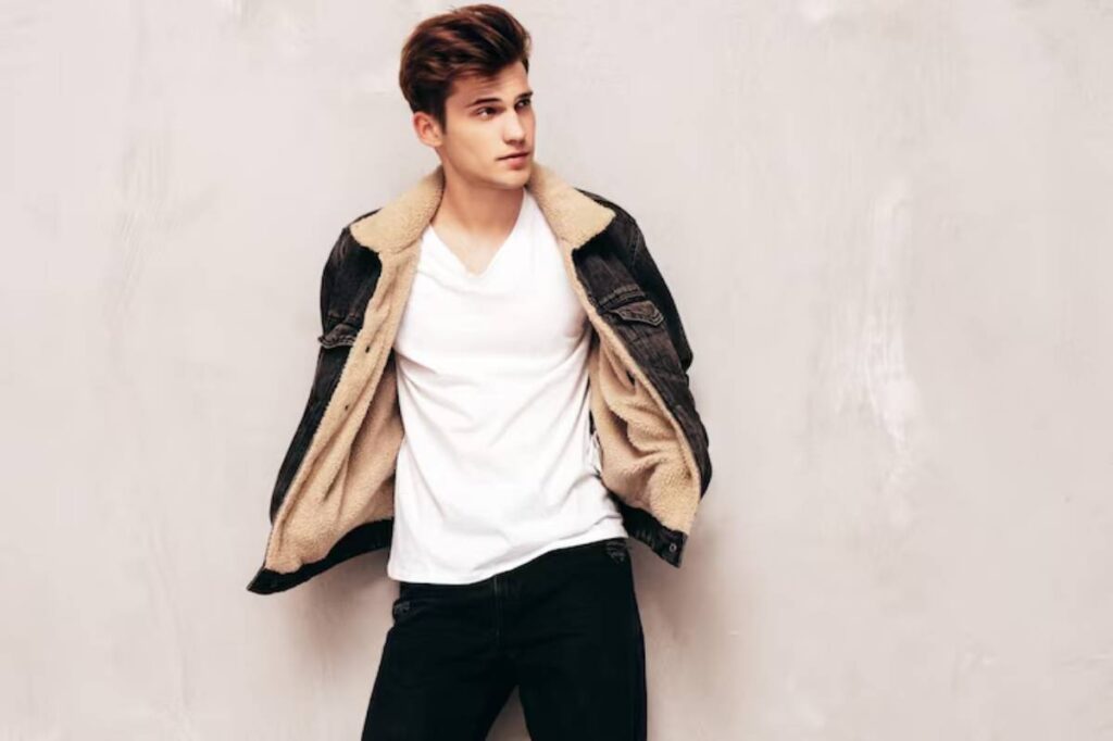 A person wears a casual outfit with a white V-neck shirt and a dark denim jacket lined with tan fur, posed against a beige background.