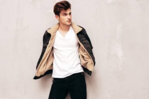 A person wears a casual outfit with a white V-neck shirt and a dark denim jacket lined with tan fur, posed against a beige background.