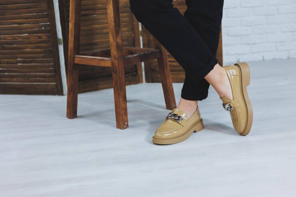 A close-up of a person’s feet in stylish tan loafers with a chain detail, paired with black pants, next to a wooden stool.