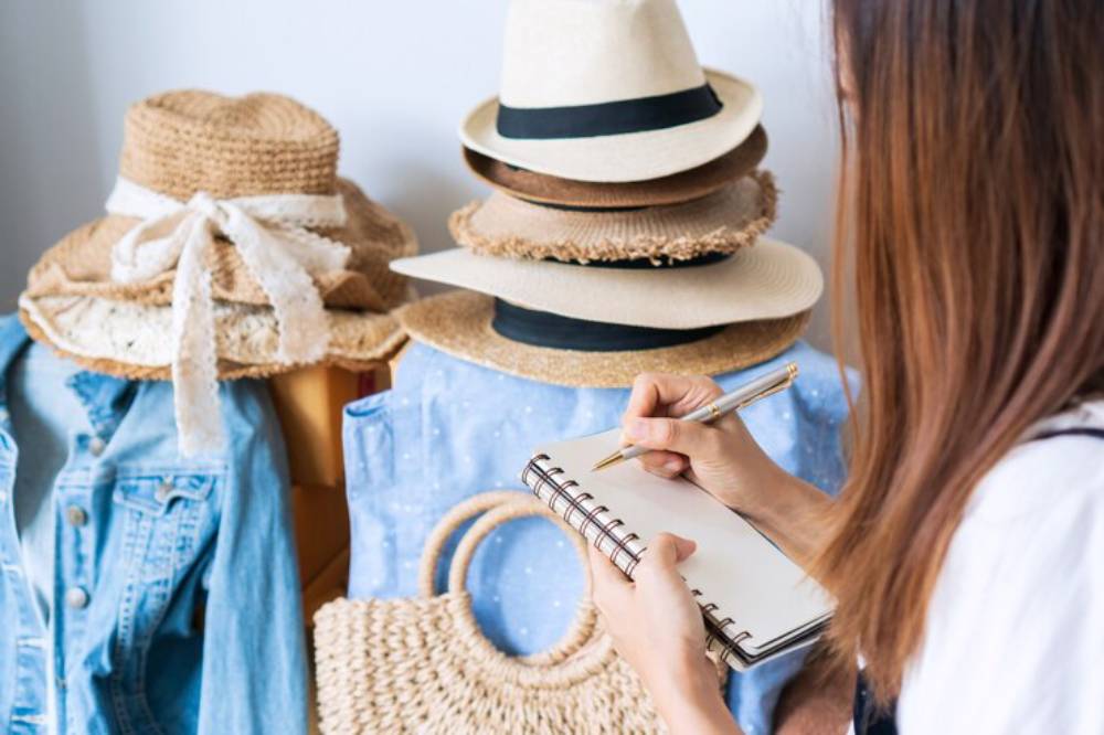 A woman writes in a notebook while surrounded by various straw hats and casual clothing laid out on a desk.