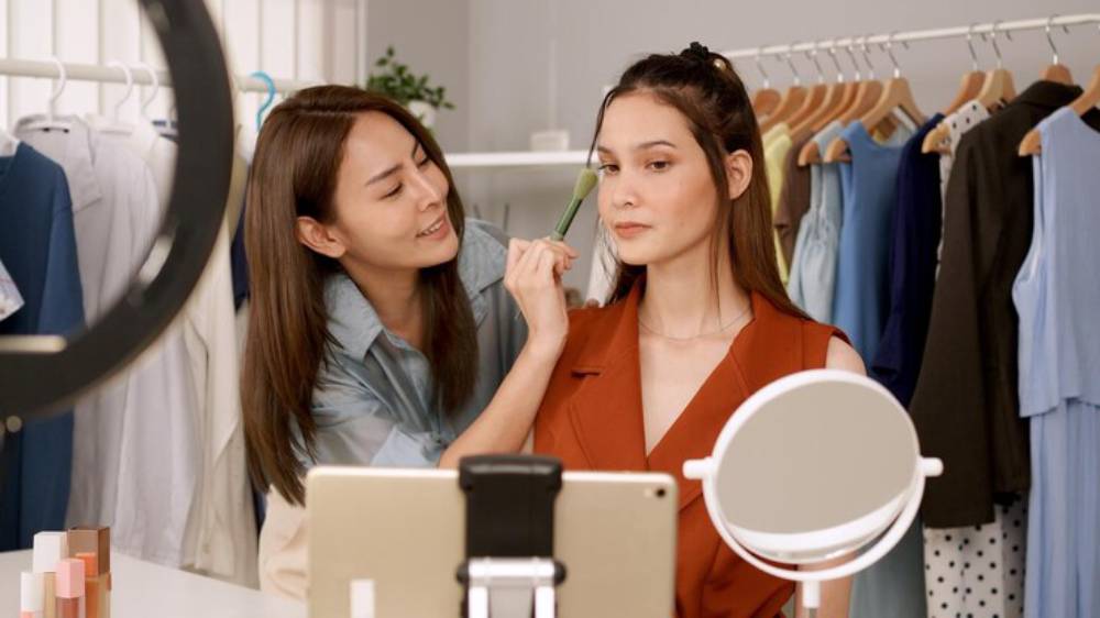 A woman applies makeup to another woman in a stylish dressing room, surrounded by colorful clothing on hangers and a vanity mirror.