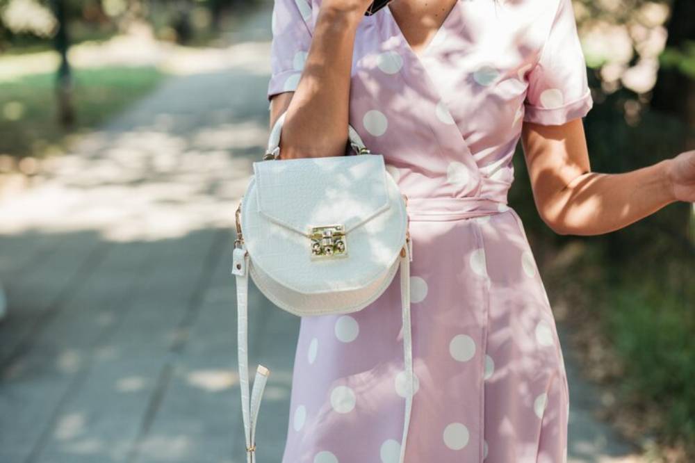 A woman in a pink polka dot dress holds a stylish white handbag while talking on her phone, standing on a sunlit pathway.