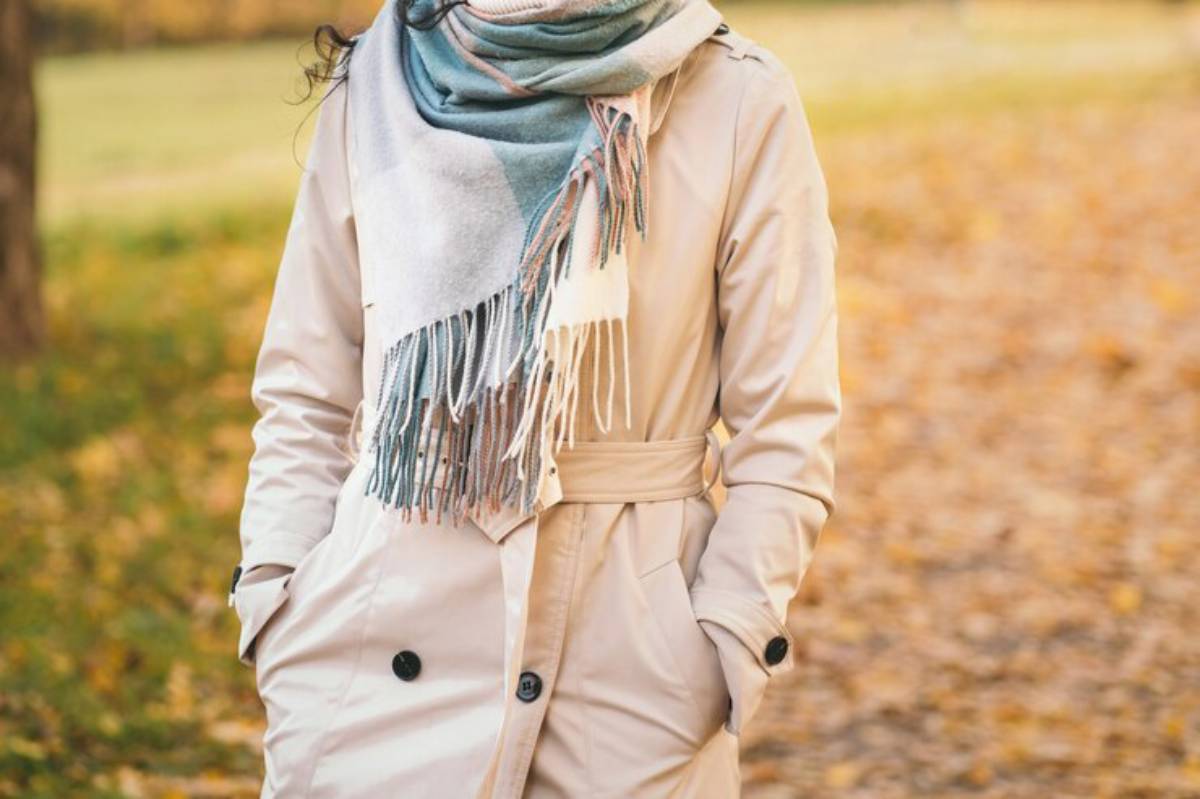 A woman in a beige trench coat and a colorful scarf stands amidst autumn leaves, hands in pockets, enjoying the outdoors.