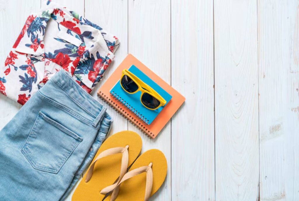 A floral shirt, denim jeans, yellow flip flops, and colorful notebooks paired with sunglasses on a wooden surface.