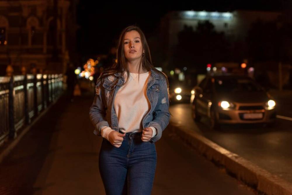 A person walks confidently along a busy street at night, wearing a denim jacket and jeans with city lights and cars in the background.