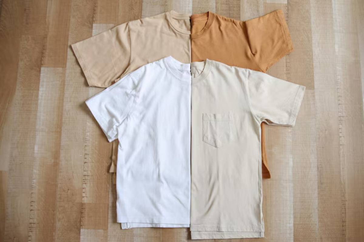 Three short-sleeve t-shirts in white, beige, and light brown arranged on a wooden surface, showcasing casual fashion options.